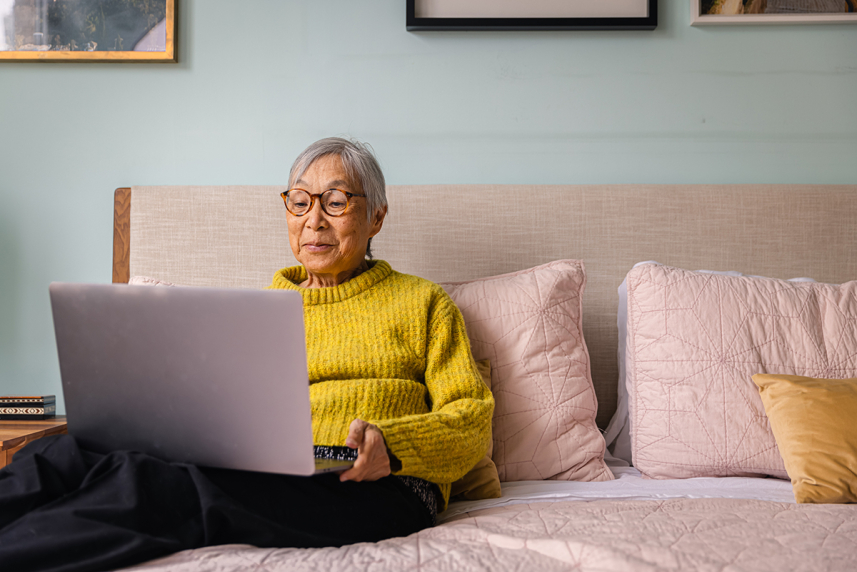 A senior sits on a bed with a laptop